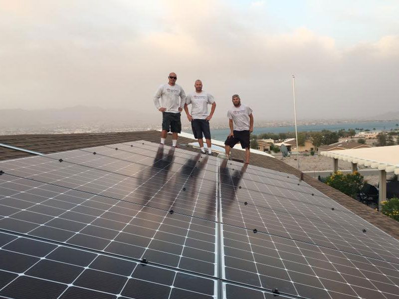 Three people standing on solar panels at sunset, overlooking a coastal landscape.