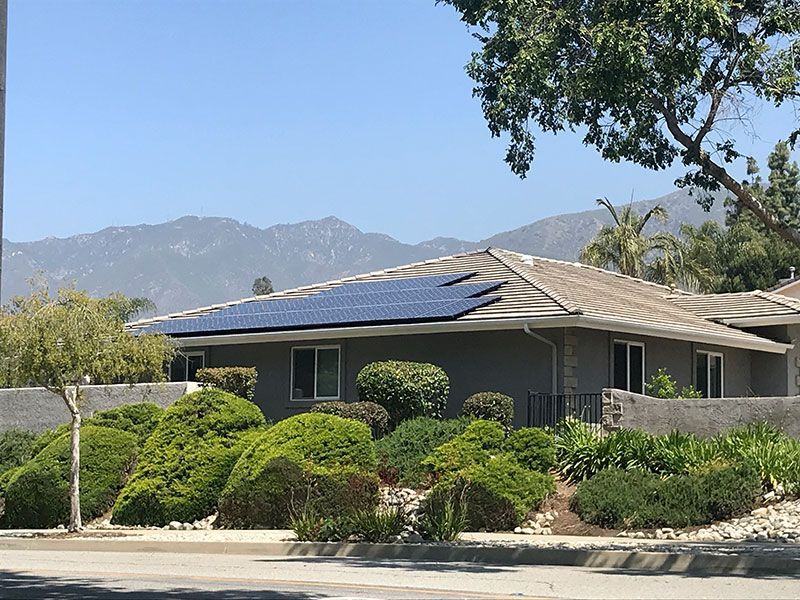 Single-story house with gray roof, green shrubs, and mountains in the background under a clear blue sky