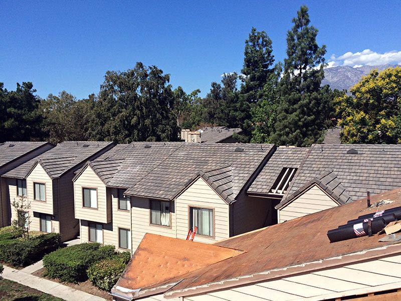Suburban apartment rooftops with tan siding and trees under a blue sky, one roof being repaired