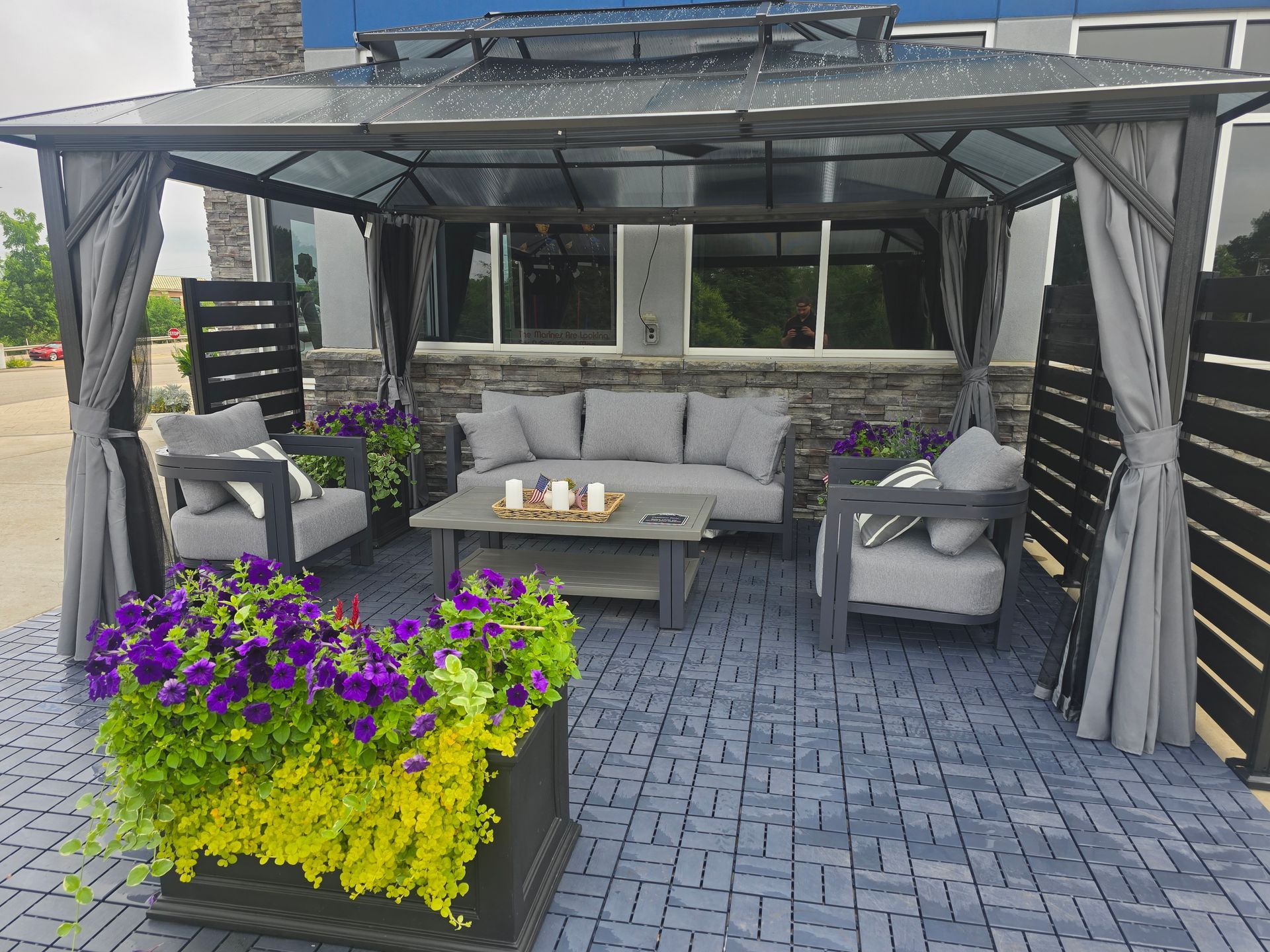 Covered patio with gray sectional seating, curtains, and purple-yellow flowers on a paved deck | Strother Brothers Auto Care