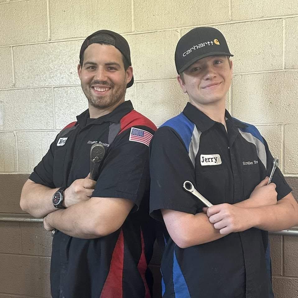 Two smiling workers in black uniforms and caps stand back-to-back with arms crossed against a beige wall | Strother Brothers Auto Care