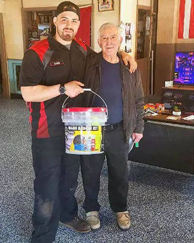 Two people standing indoors, one holding a bucket and both posing beside a TV and table | Strother Brothers Auto Care