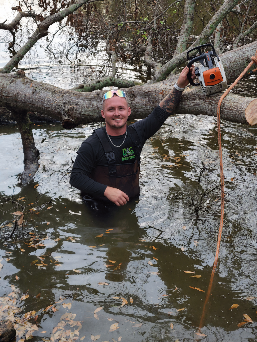 Man standing in muddy water, holding a chainsaw beside a fallen tree over a creek.