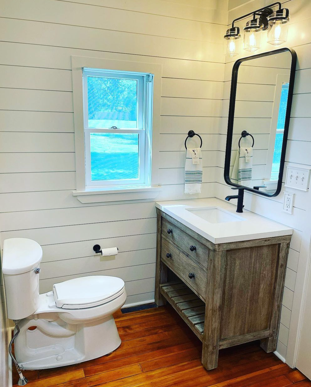 A bathroom with a toilet , sink , mirror and window.
