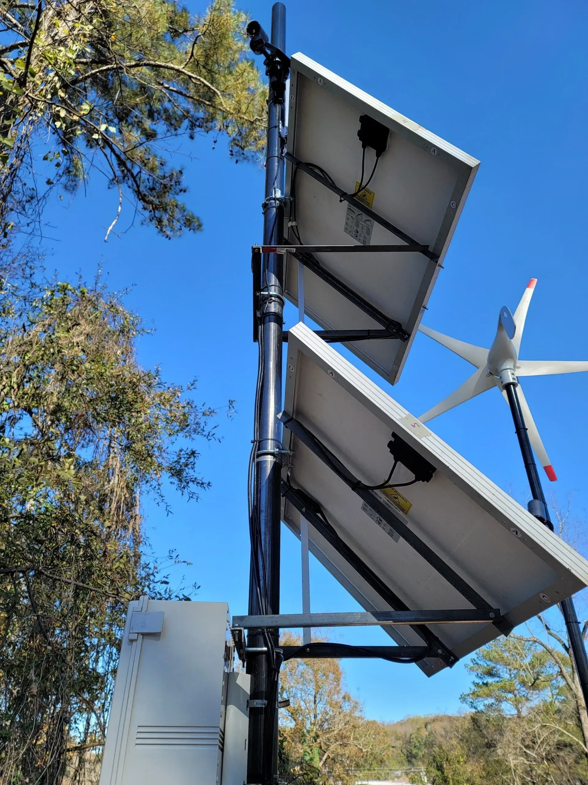 Solar panels mounted on a pole, angled toward the sky, with a small wind turbine in the background against a blue sky.