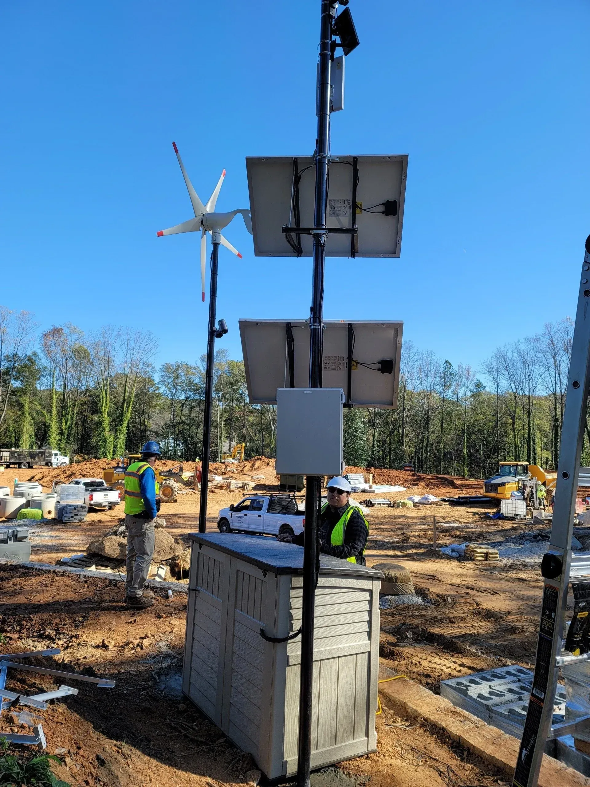Construction site with workers, a wind turbine, solar panels, and a utility box. Blue sky in the background.