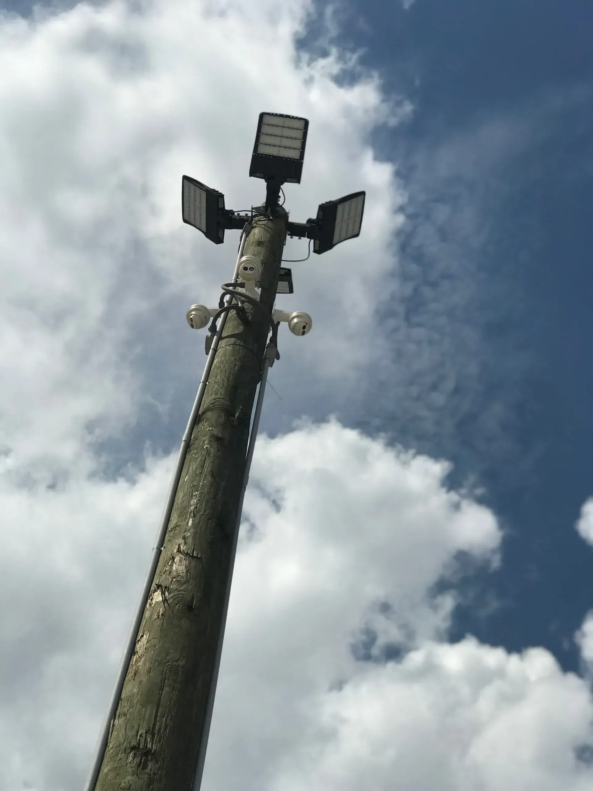 A tall wooden utility pole with bright LED floodlights and surveillance cameras against a cloudy blue sky.