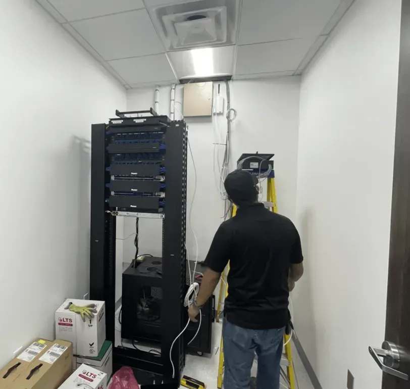 Man working in a server room with black server rack and ladder. Wires and equipment fill the small, white-walled space.
