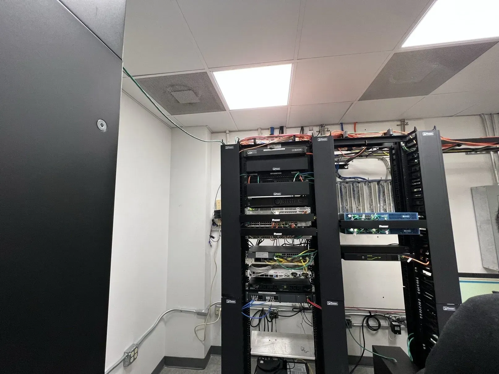 A server room with black racks filled with electronic equipment, wires, and cables. The room has a white ceiling and walls.