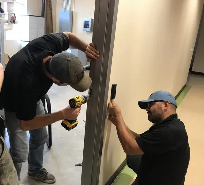 Two men installing a door frame in a room with beige walls. One uses a power drill, the other holds a piece of the frame.