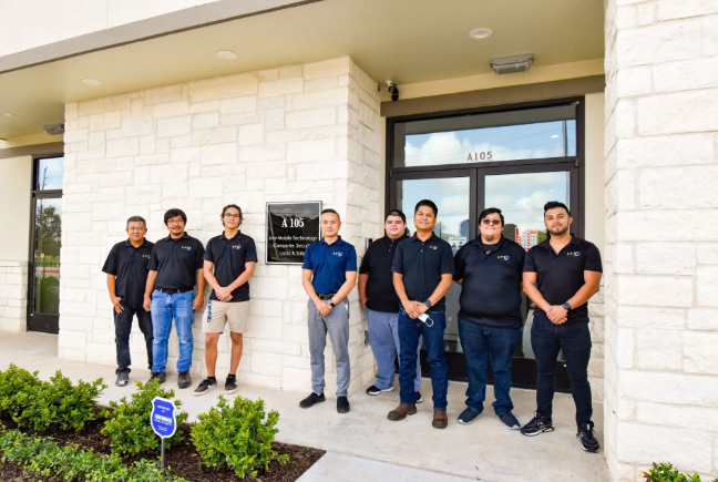 Group of men in matching shirts standing outside a building with a sign. They pose for a photo in front of a stone facade.