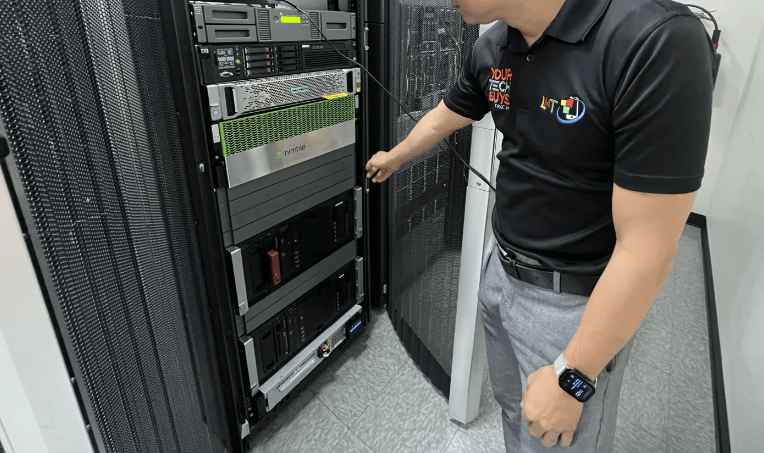 A man in a black polo shirt opens a server rack door in a data center, pointing towards the equipment inside.