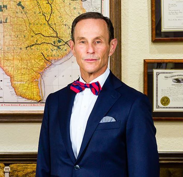 Man in a blue suit and bow tie stands in front of a map and framed documents.