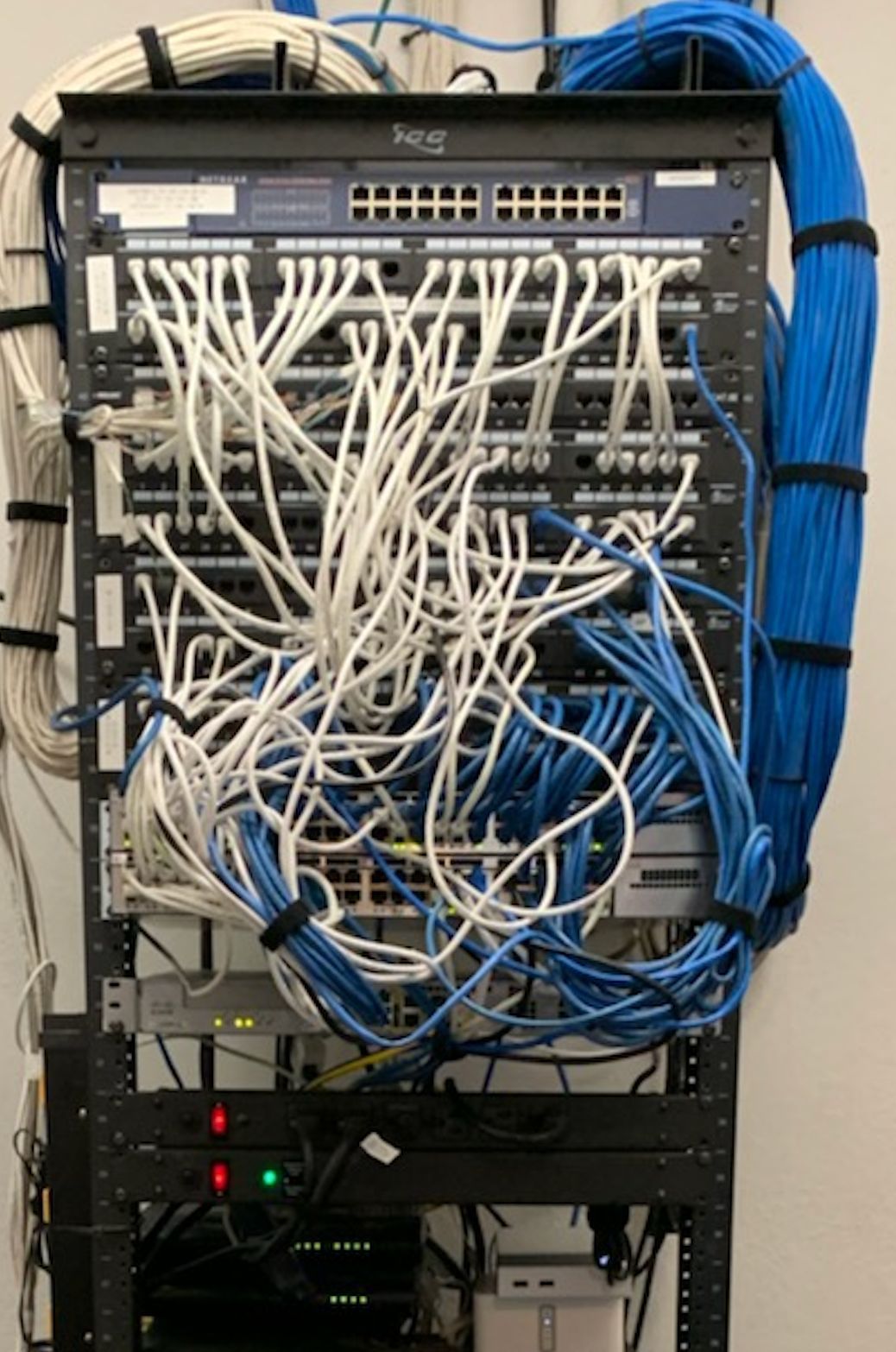 A messy network rack with numerous tangled white and blue network cables connected to switches. Cables are bound in some areas with black velcro straps.