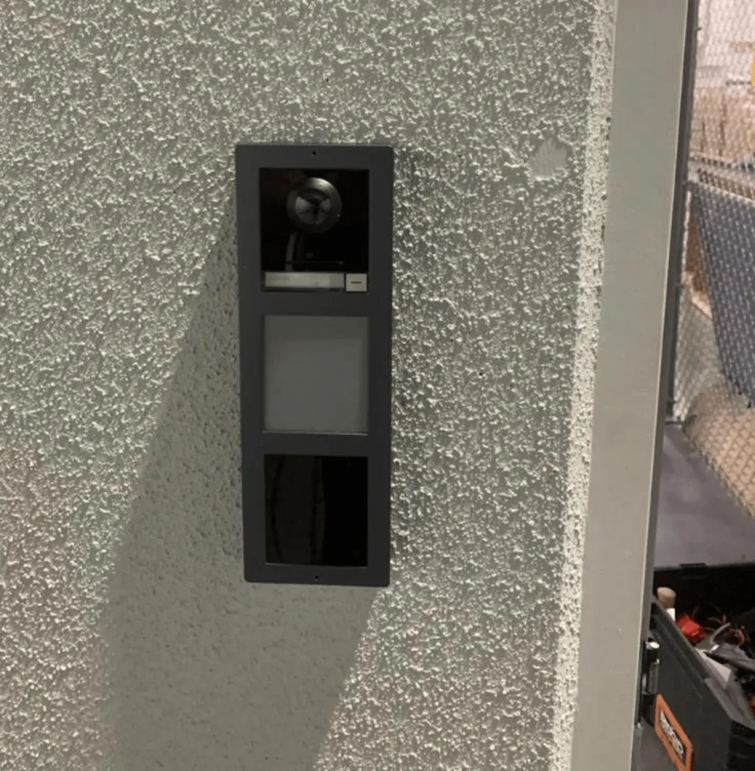 A dark gray doorbell unit with a camera and buttons is mounted on a textured, white stucco wall next to a white pillar.