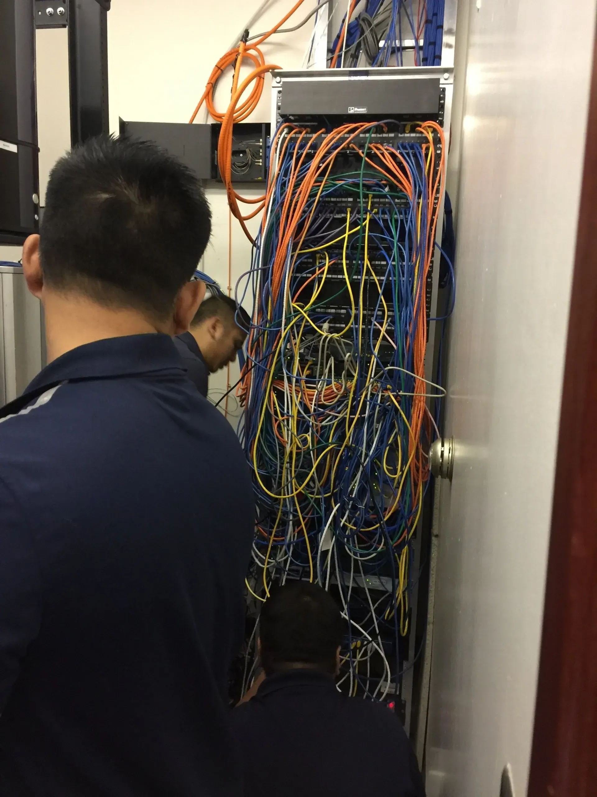Two men work on a tangled network server rack filled with colorful cables in a small room.