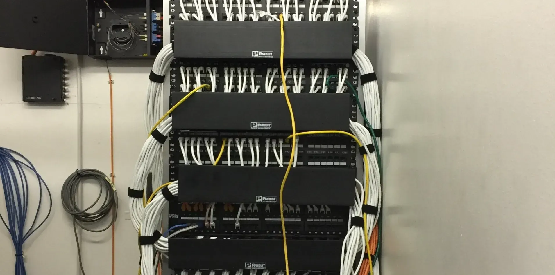 Network rack with black components, numerous white and yellow network cables, installed against a light-colored wall.