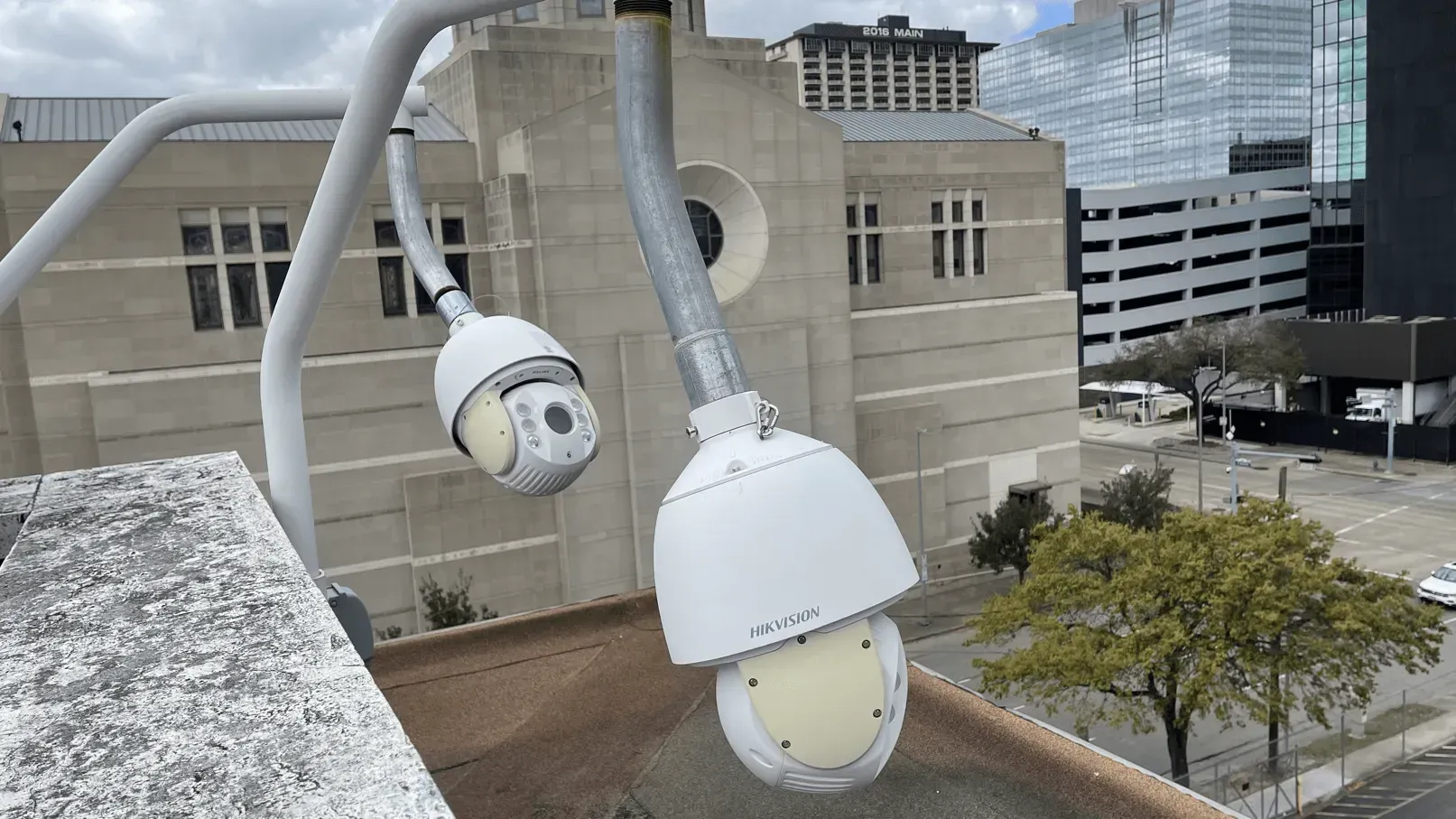 Two white surveillance cameras hanging from a curved metal arm on a rooftop. A building and trees are in the background.