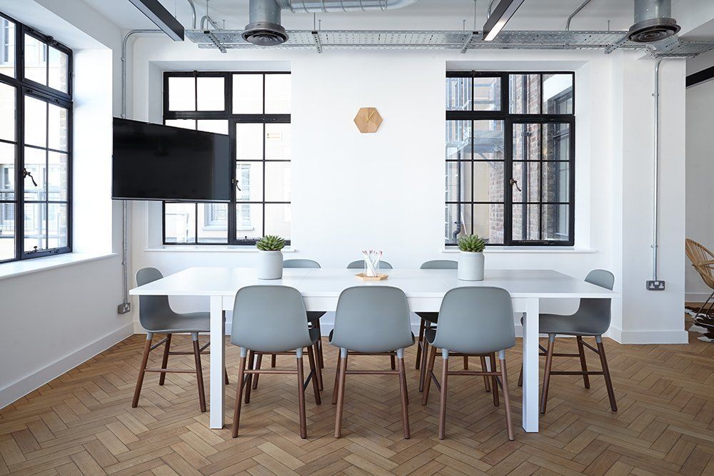 A bright meeting room with a long white table, gray chairs, and large windows; a mounted TV and clock hang on the white wall.