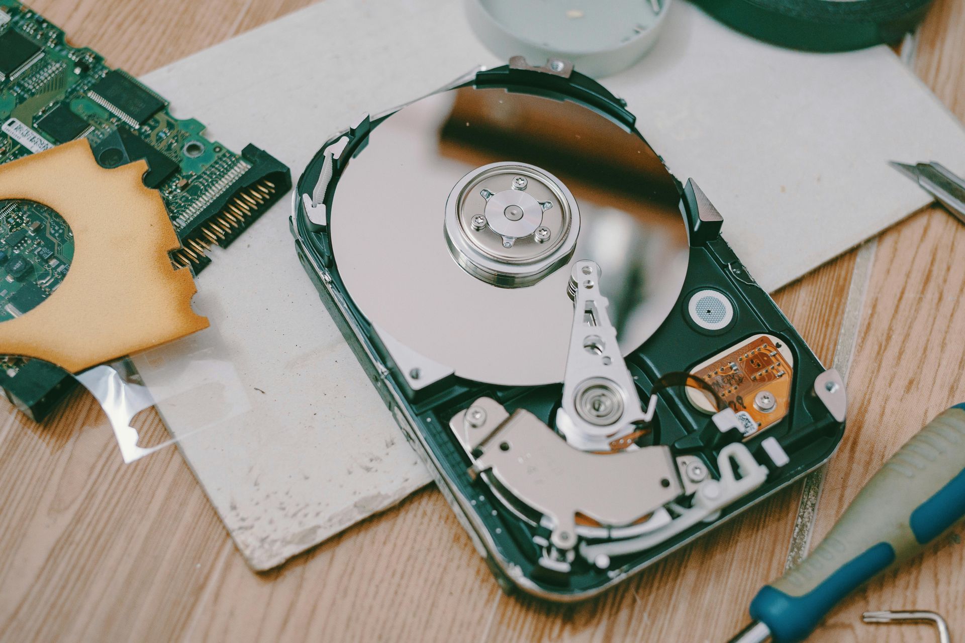 Open hard drive with exposed platters and internal components, alongside electronic parts and tools on a wooden surface.