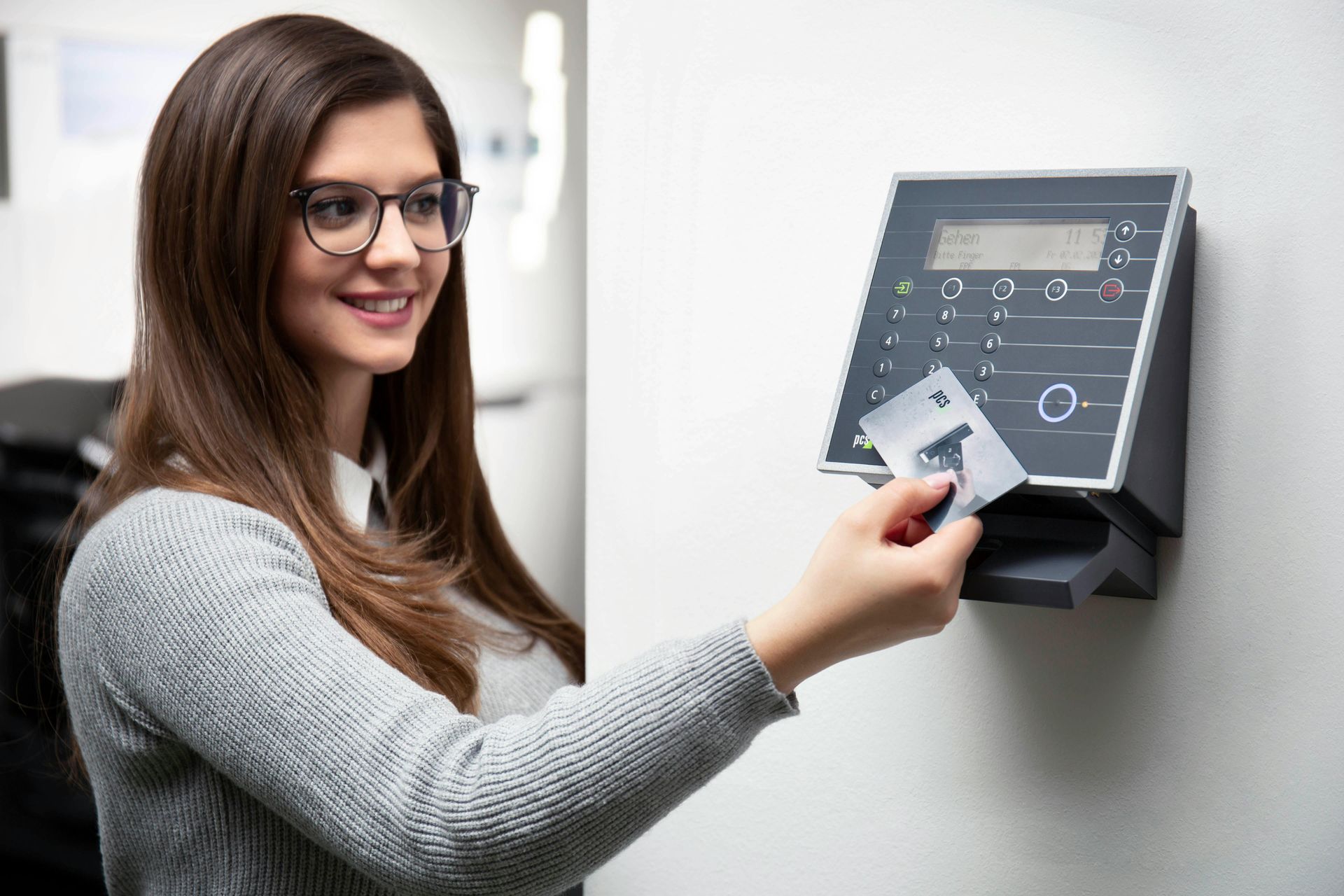 A woman with glasses scans her card at a time clock in a white-walled office. She smiles.