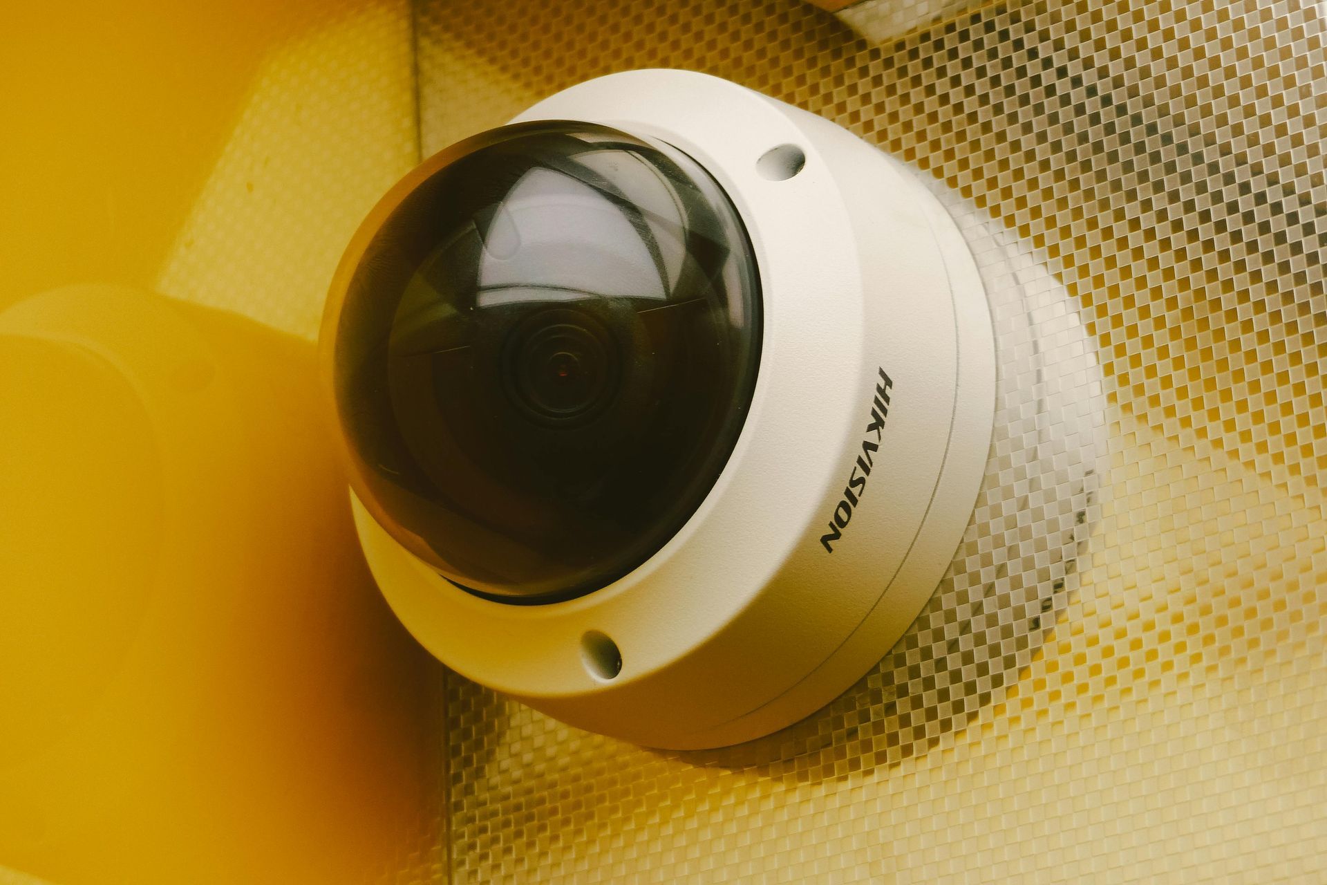 Security camera, white and dark dome, mounted on a textured surface next to a yellow wall.