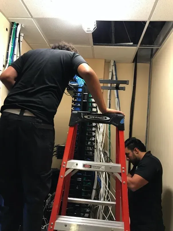 Two men working on network cables in a server room. One on a ladder, the other by the server rack. Ceiling tiles are missing.