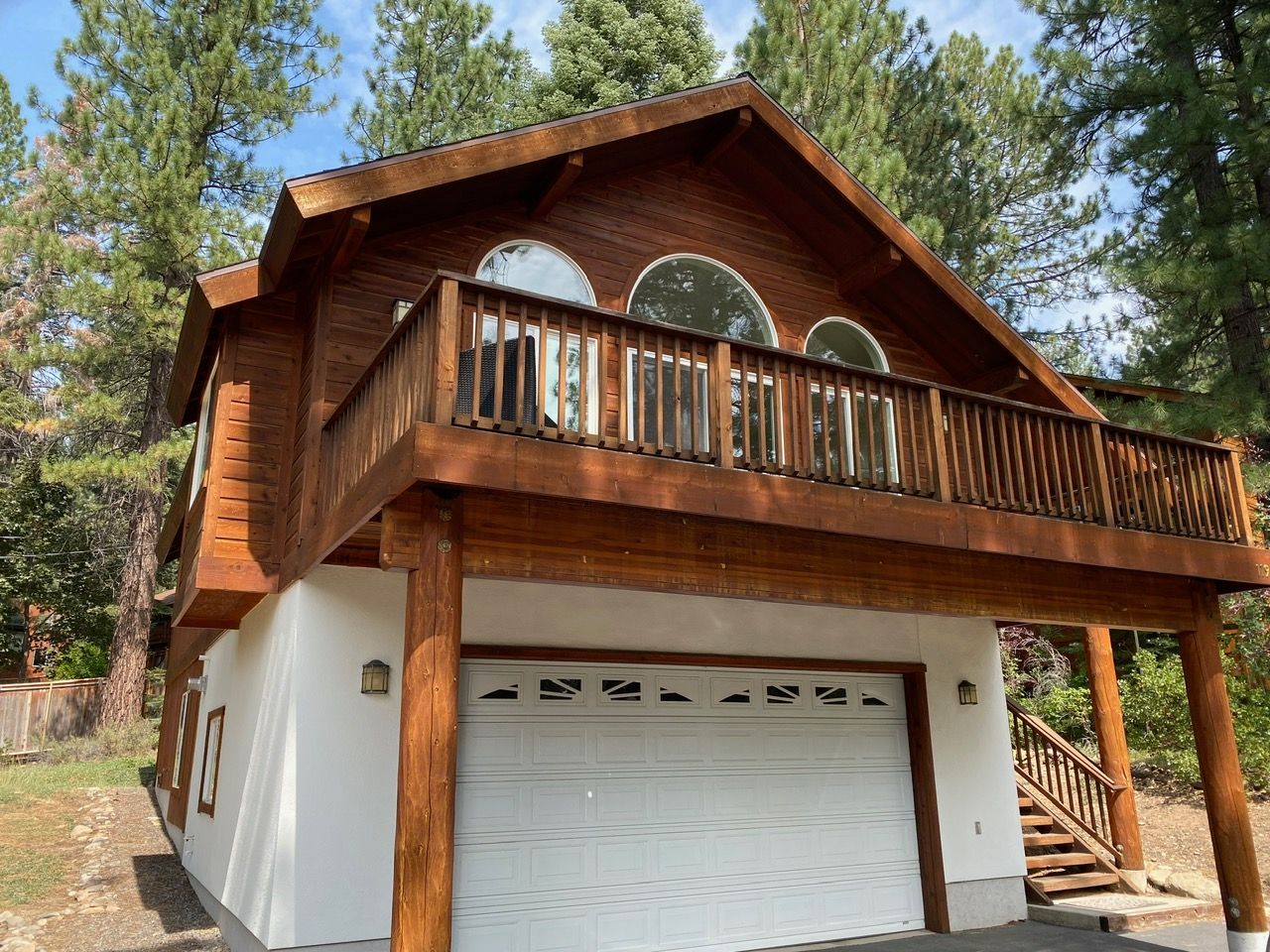 A rustic two-story cabin with white garage door, wooden balcony, and pine trees surrounding it.