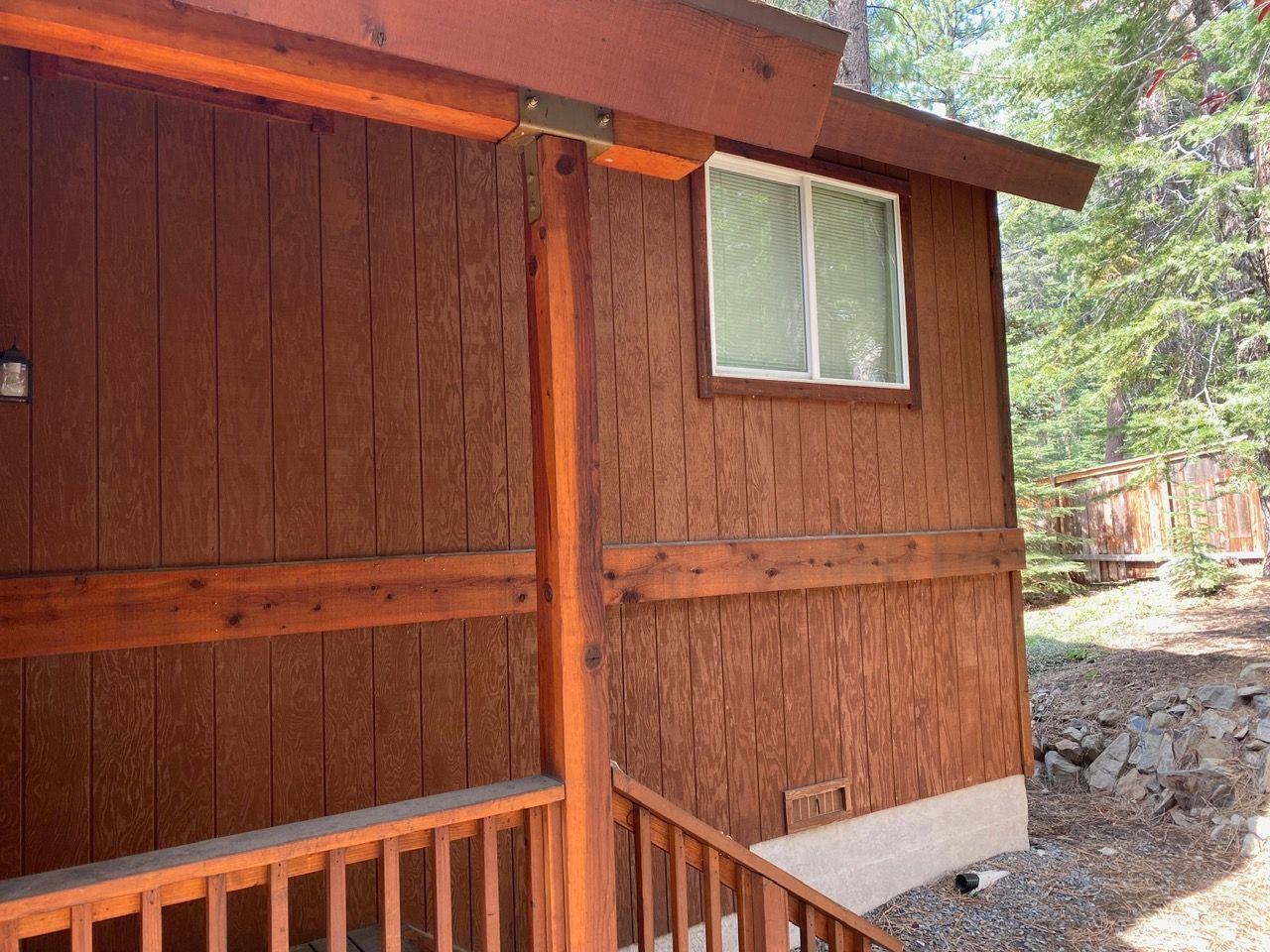 Wooden cabin exterior with a small window, porch railing, and rocky ground beside trees