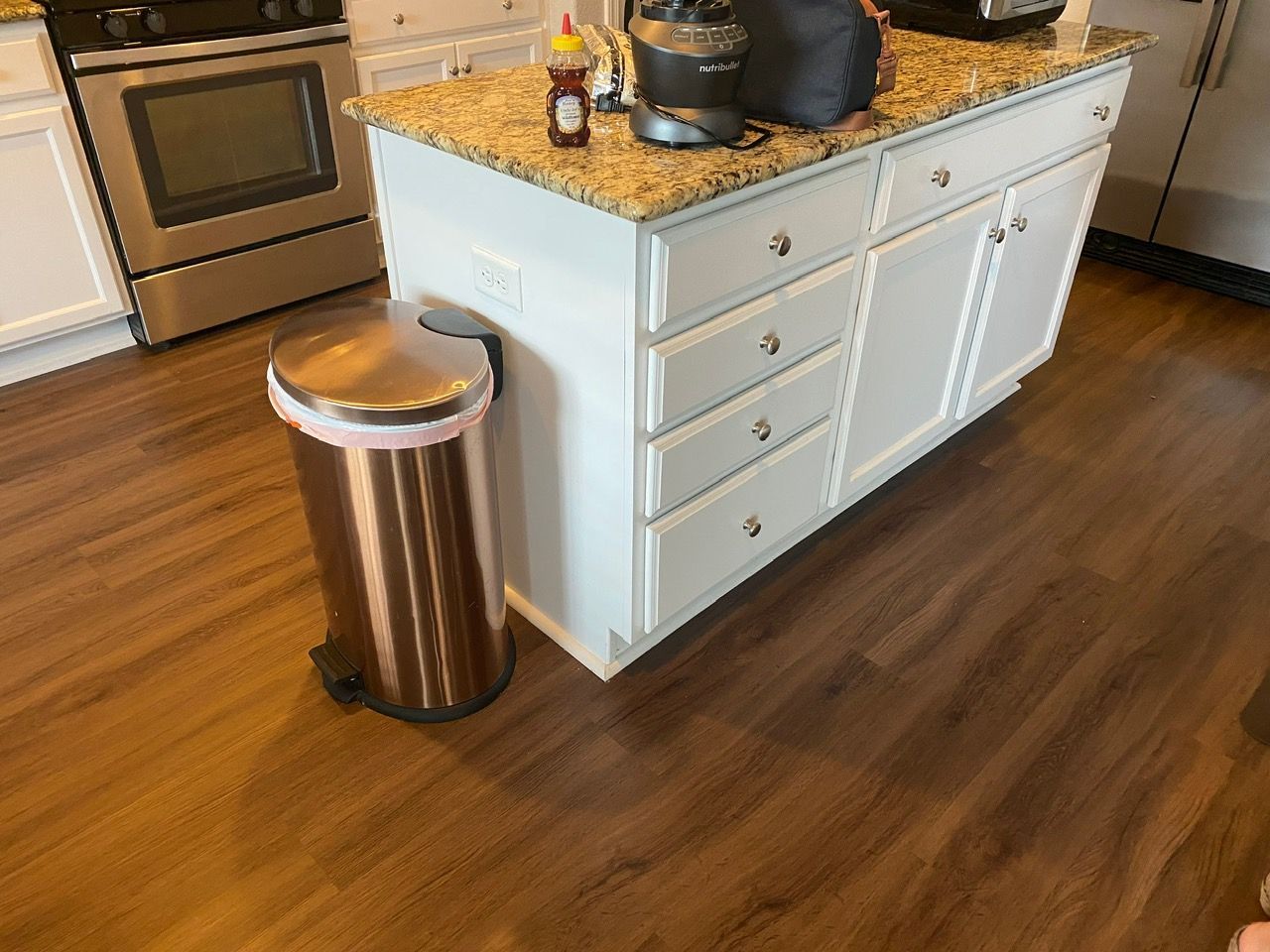 Kitchen island with granite countertop and a stainless trash can on a wooden floor.