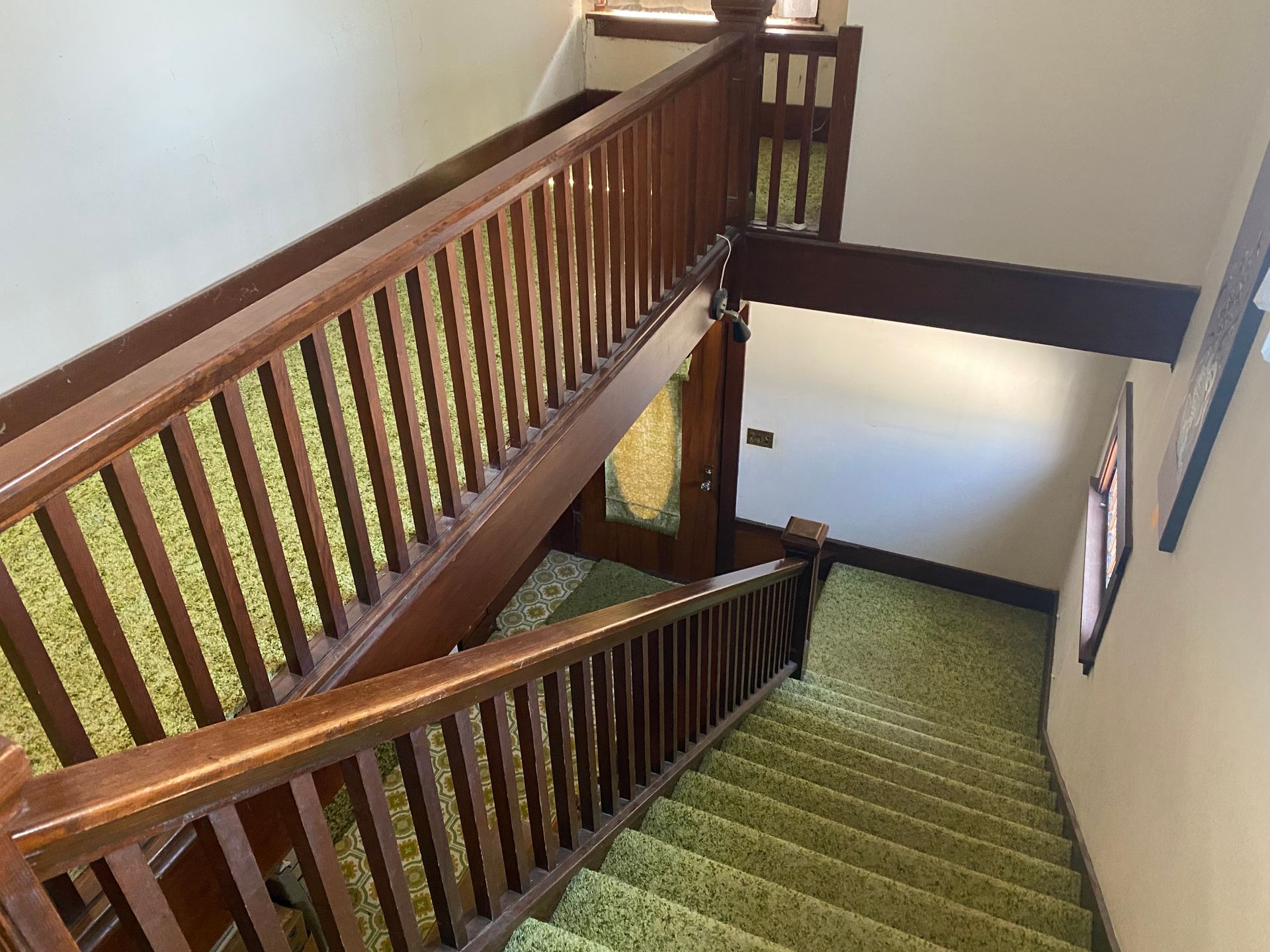 Wooden stairwell with green carpet and railings, viewed from the top landing.