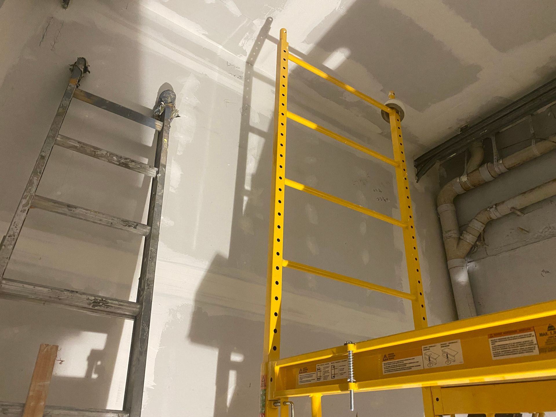 Two ladders leaning in a drywall room, one gray and one yellow, with a wooden scaffold platform in front.