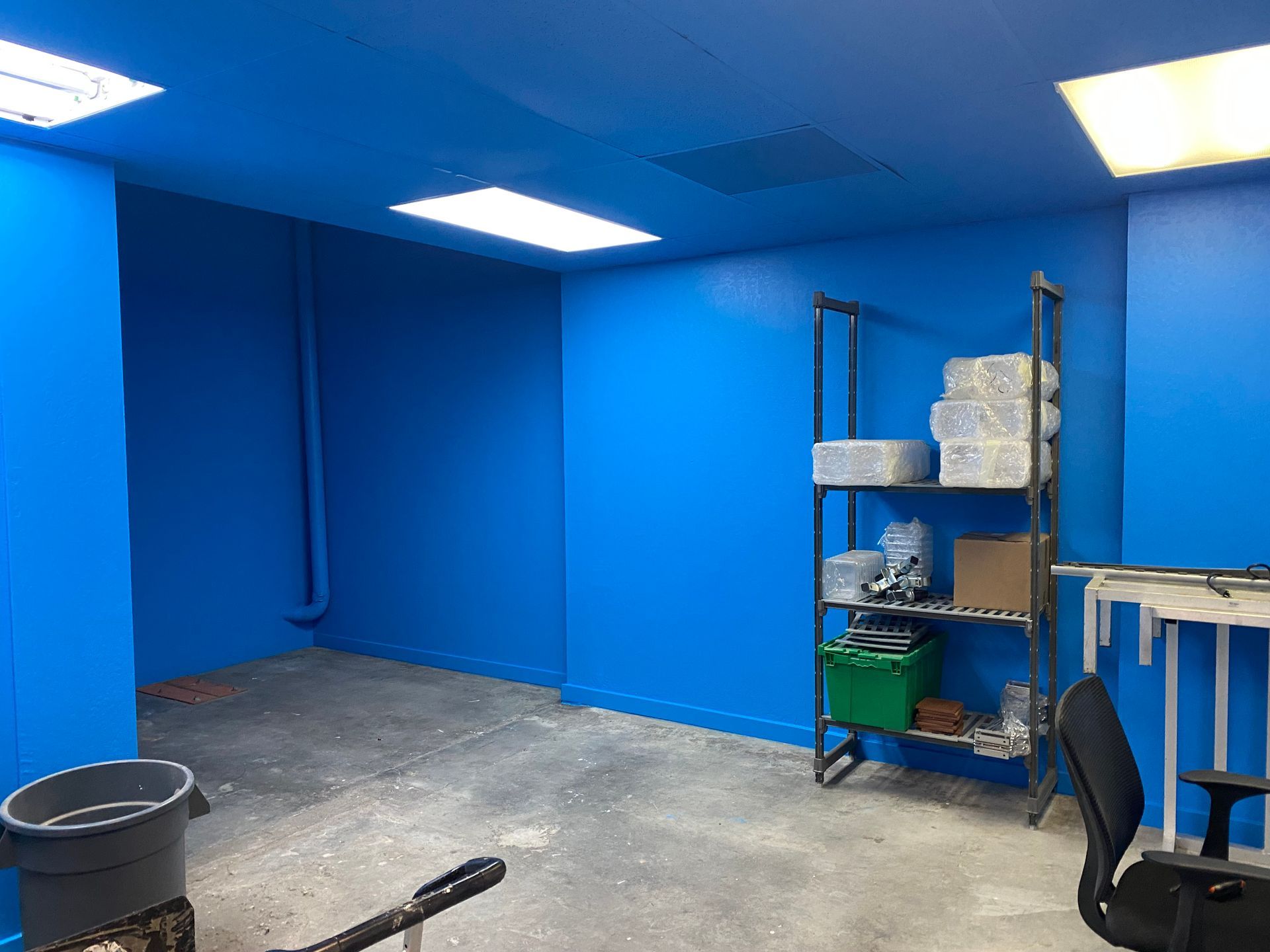 Empty blue room with gray floor, metal shelf, buckets, and a chair near a workbench.