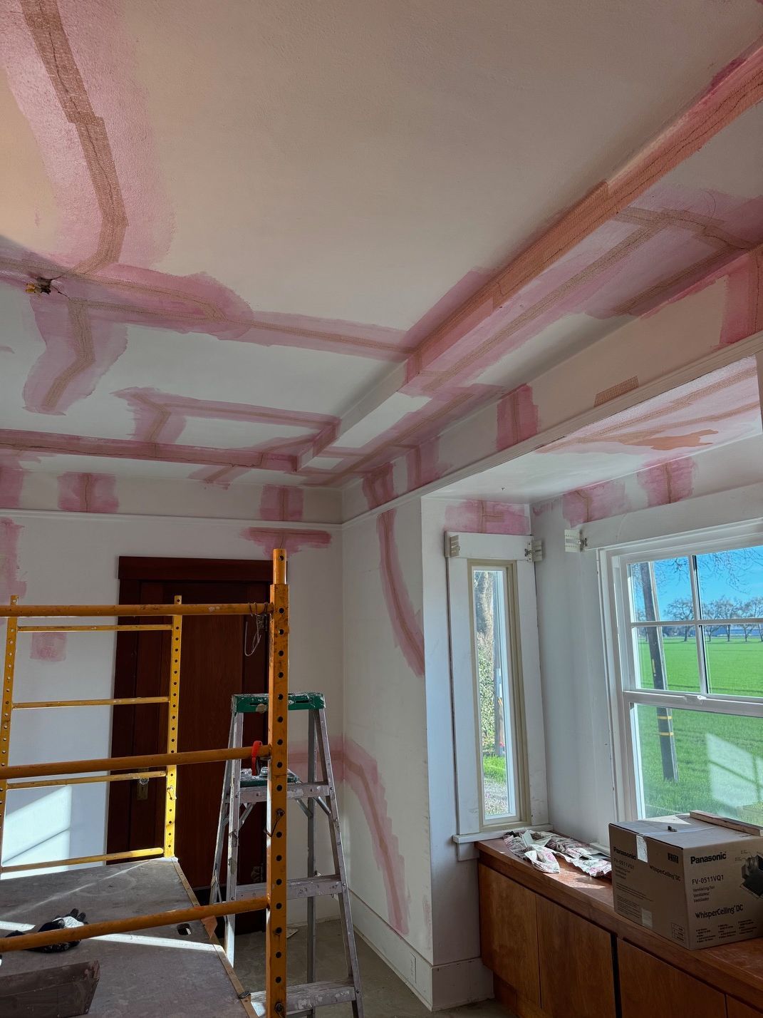 Interior room under renovation with pink drywall seams, scaffolding, and a window on the right.