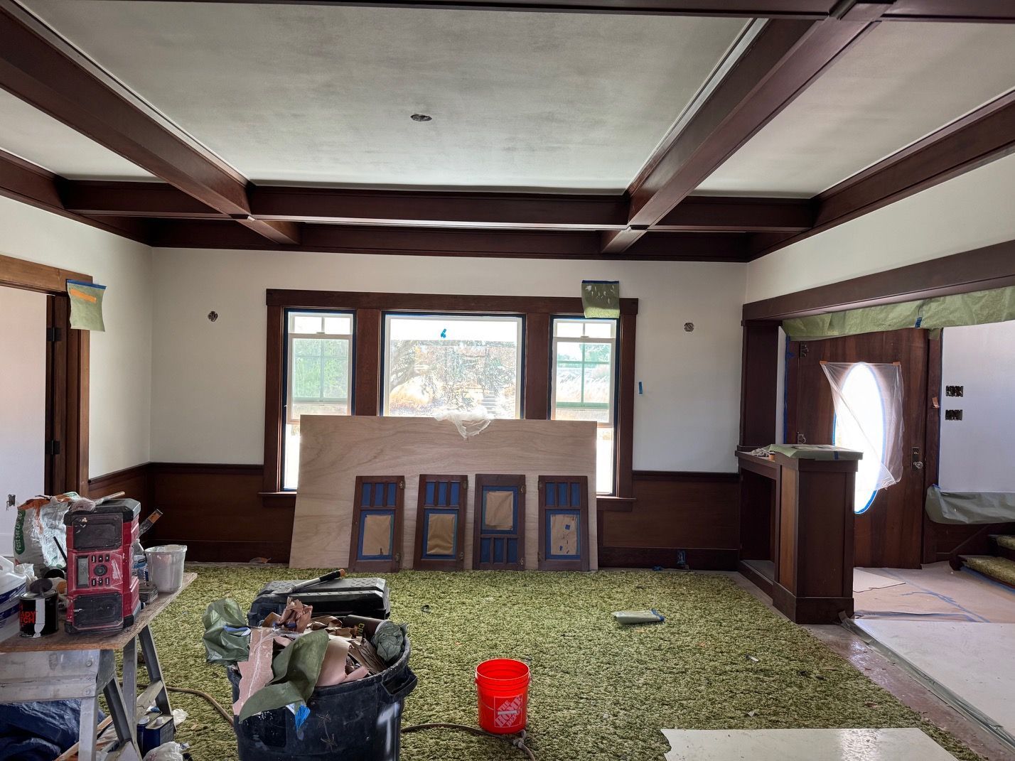 Empty living room with exposed beams, green carpet, fireplace, and a red bucket on the floor