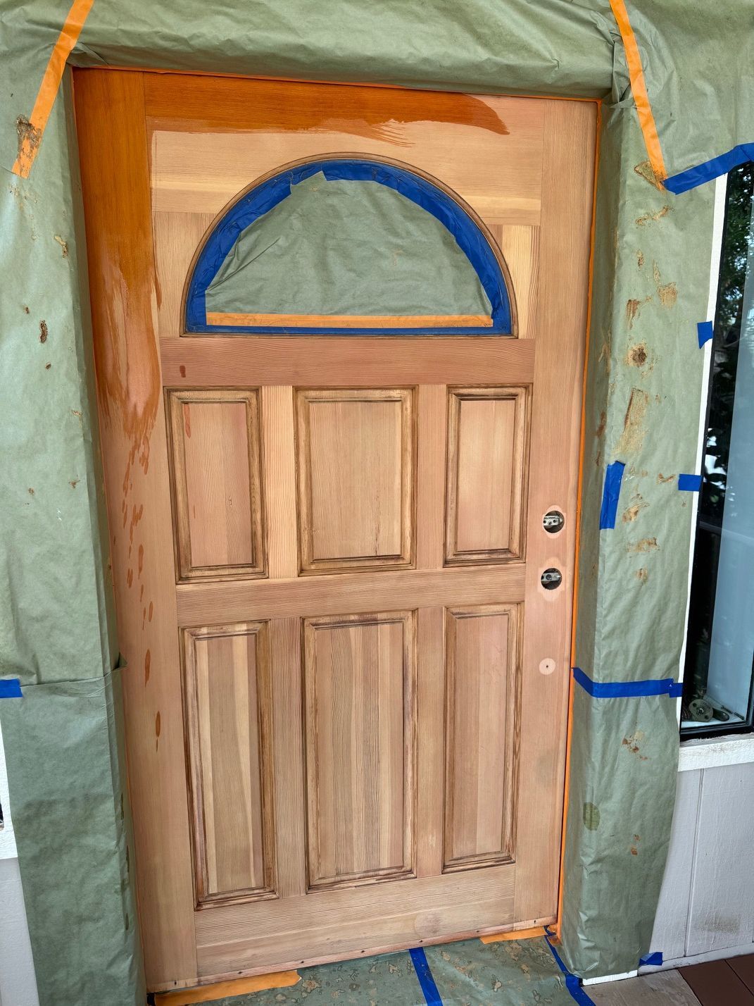 Wooden front door with arched blue-trimmed window, framed by orange trim and green protective covering