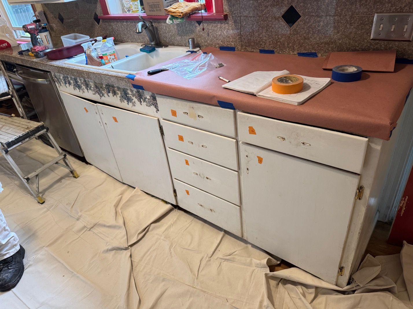 Cluttered kitchen counter with sink, white cabinets, and papers on a pink countertop over a covered floor