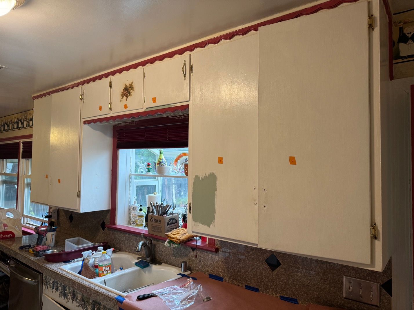 Kitchen with long white cabinets, red trim, and a window above a sink and cluttered counter.