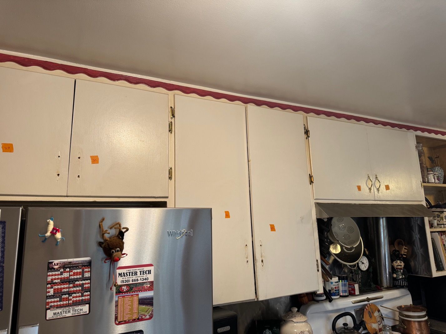 White kitchen cabinets above a stainless fridge, with red trim near the ceiling and a cluttered countertop.