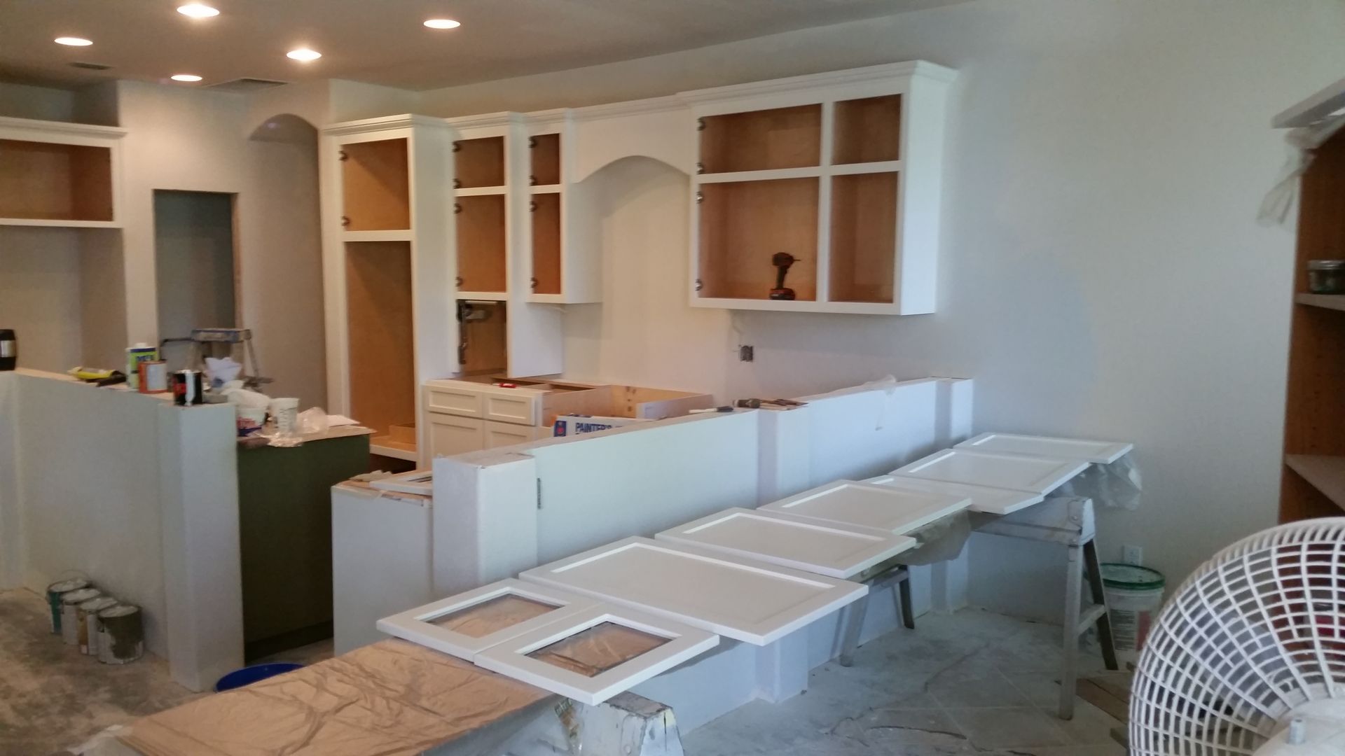 Unfinished kitchen with white cabinets, long countertop panels, and a wicker chair in a bright room