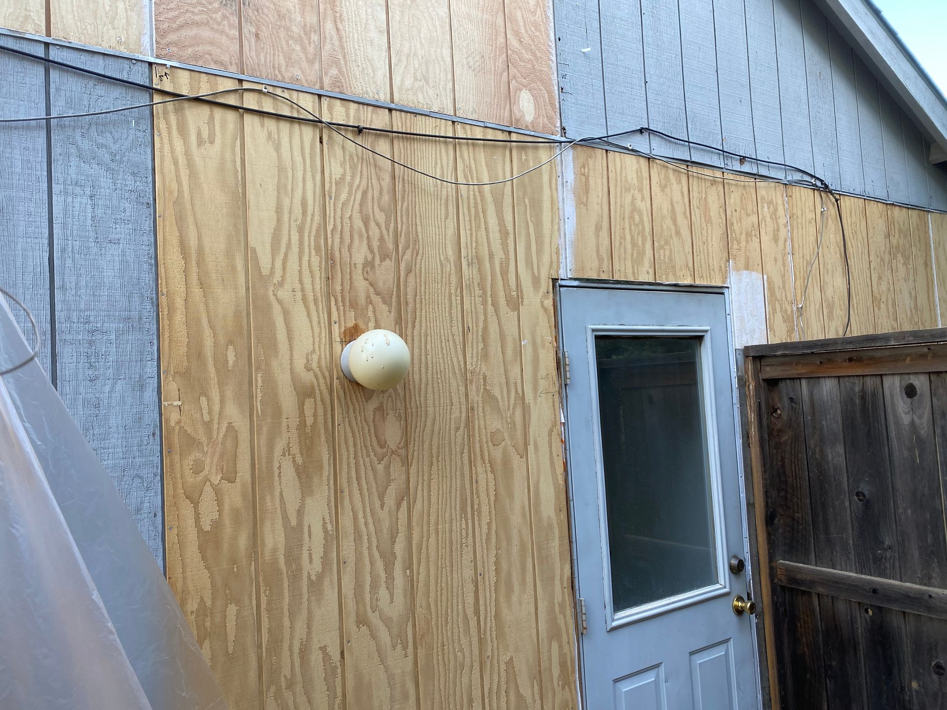 Plywood exterior wall with a white door, small window, and wall-mounted light