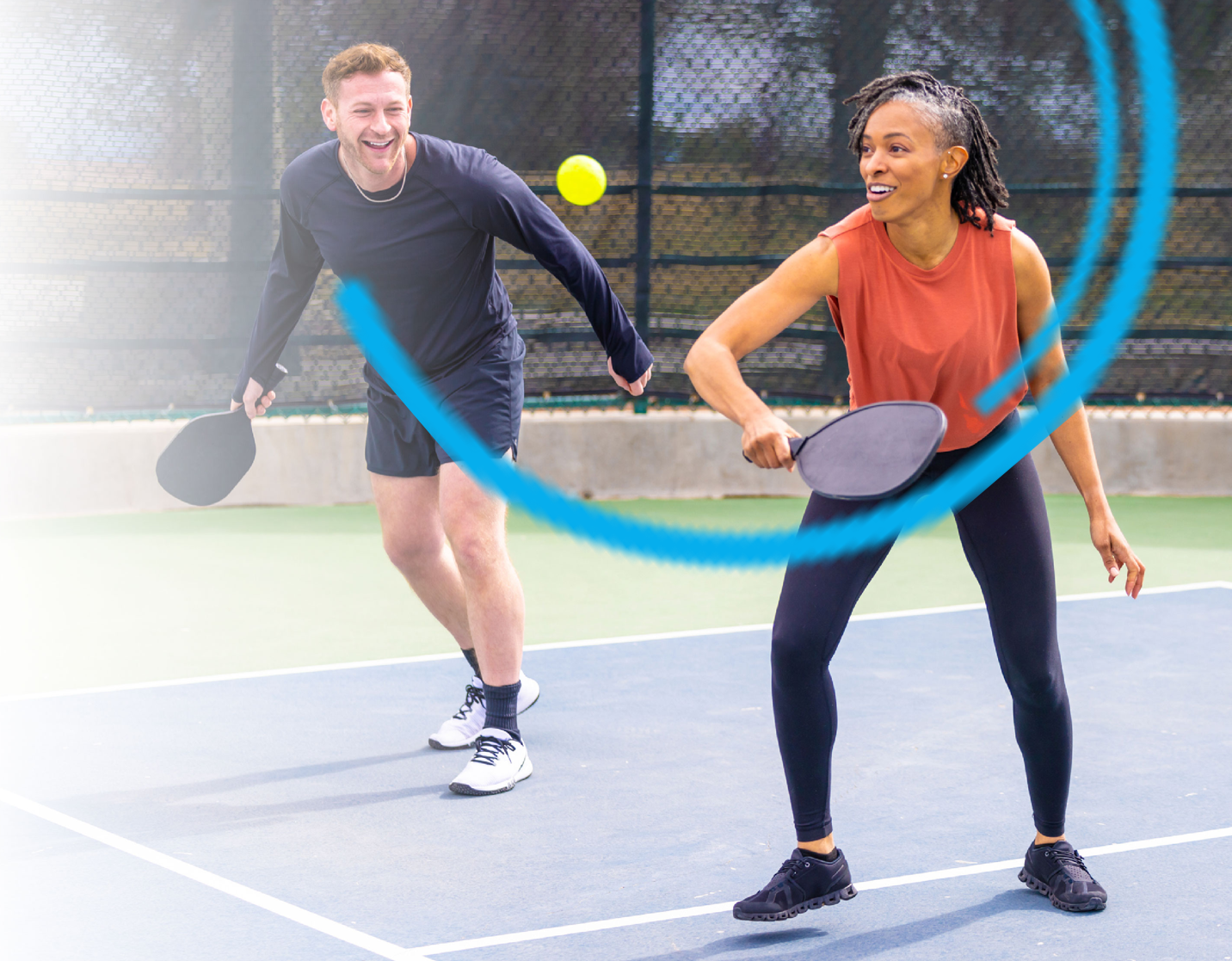 Two people playing pickleball on a court. Woman swings paddle, ball in air, man smiling with paddle.