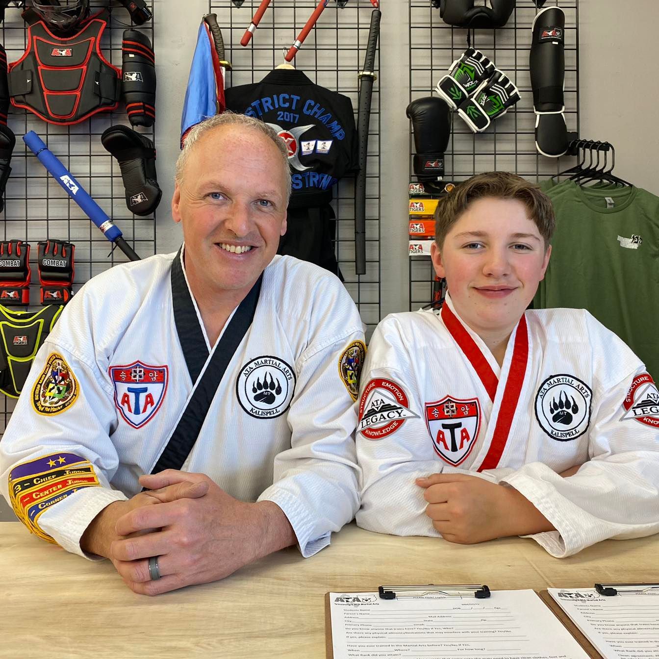Man and boy in white martial arts uniforms pose in front of equipment.