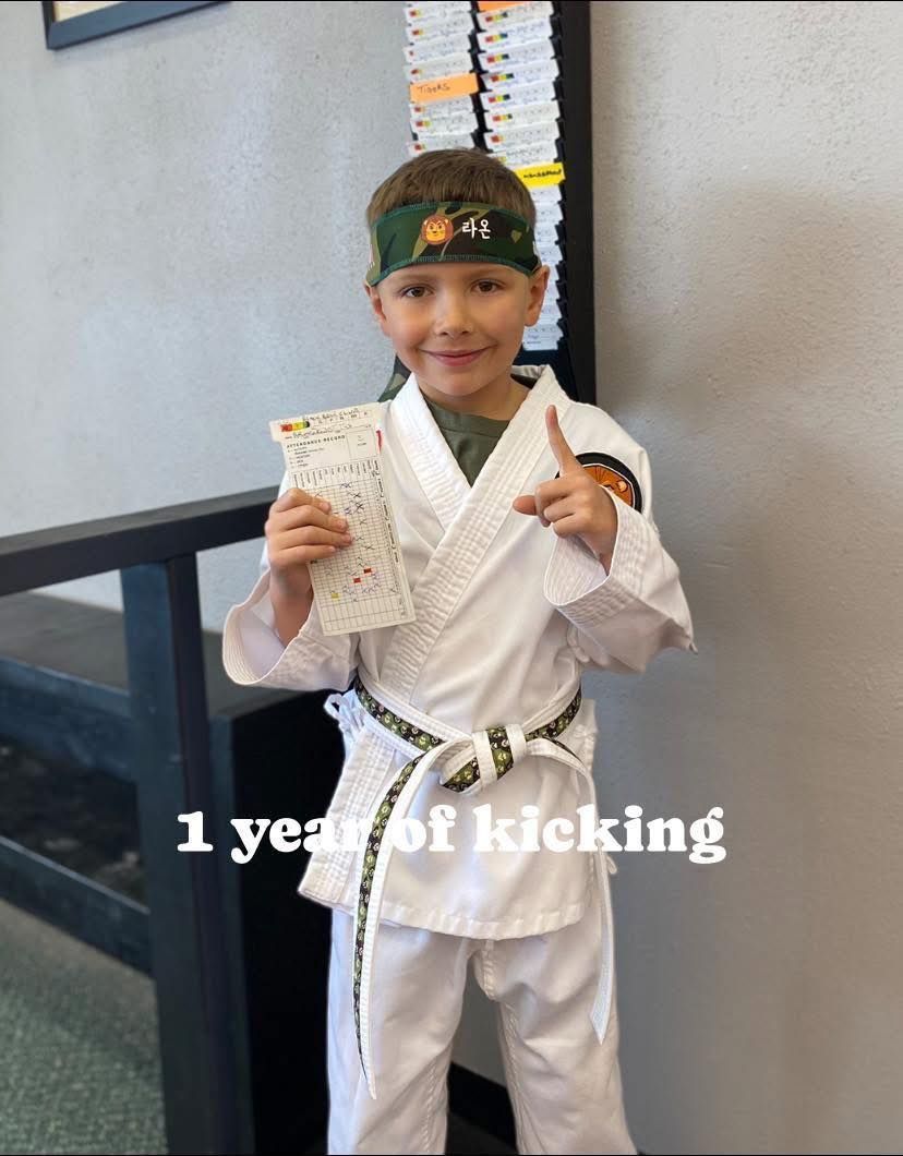 Young boy in karate uniform, holding a certificate, celebrating 1 year of kicking, smiling, pointing.