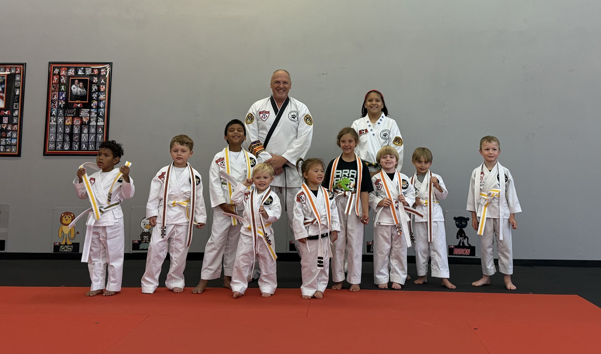 Group of young children in karate uniforms with medals, standing on a red mat with instructors.