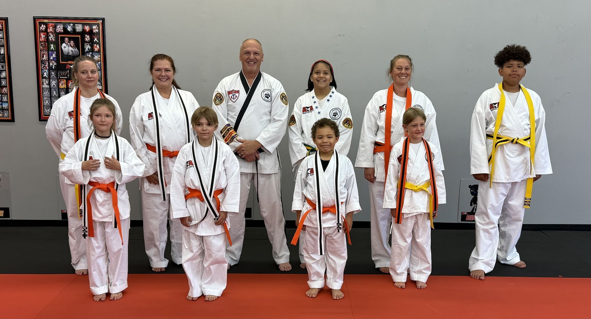 Group of martial artists in white uniforms with belts of various colors, standing on a red floor.