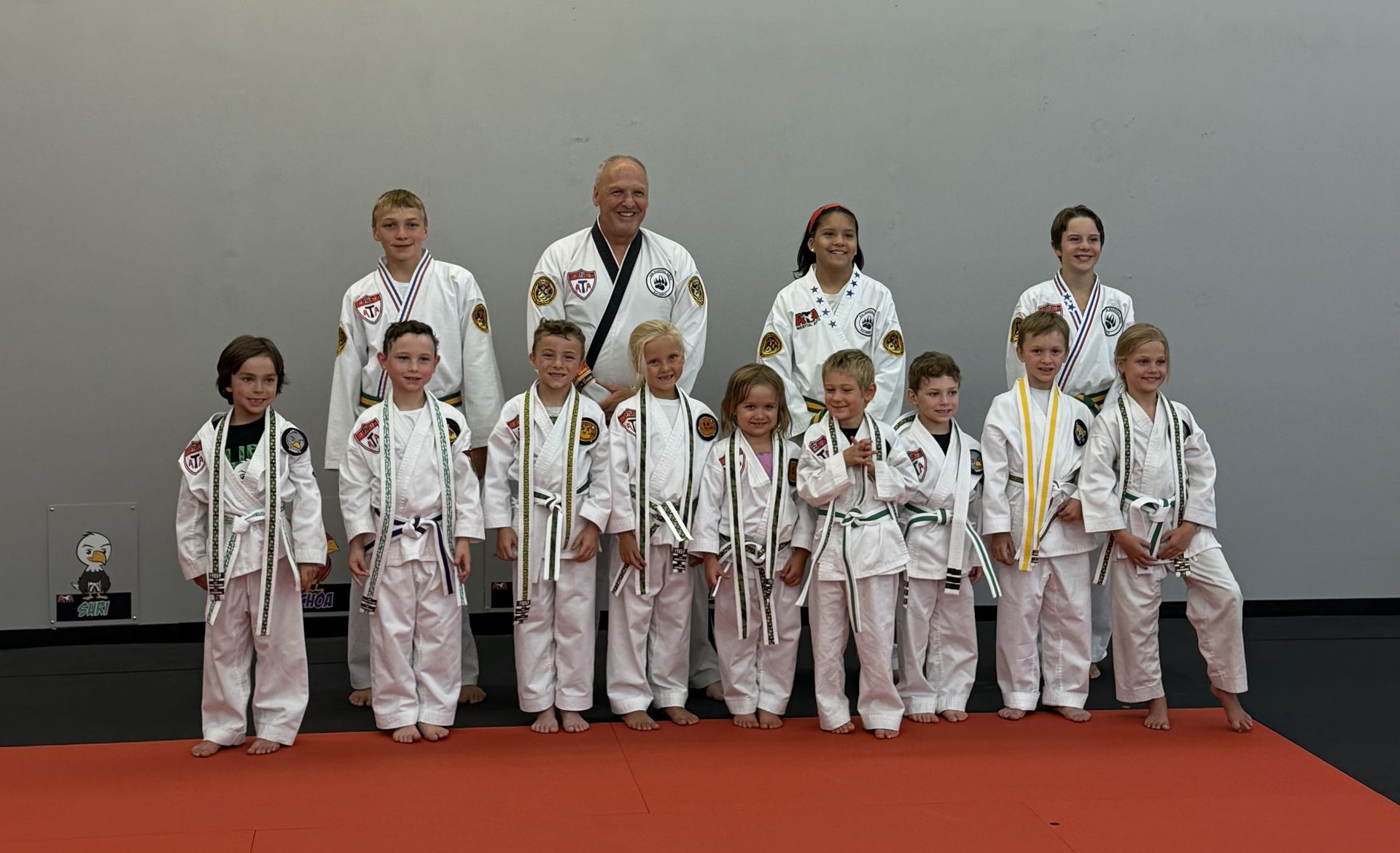 Group of children and adults in martial arts uniforms posing on a red mat.