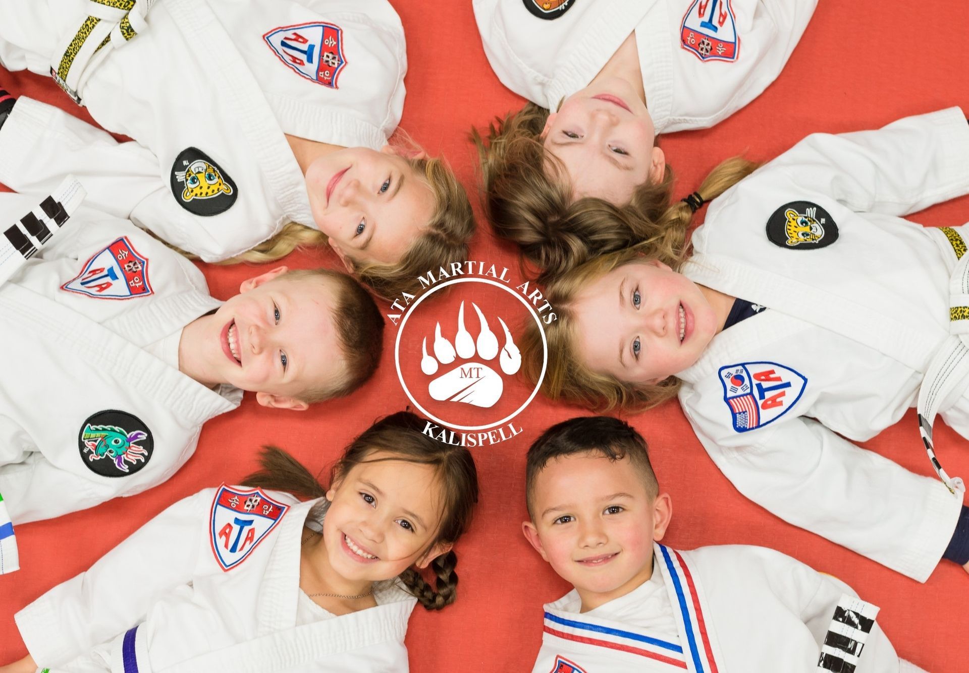 Six smiling children in karate uniforms lying in a circle on a red mat, centered on a logo.