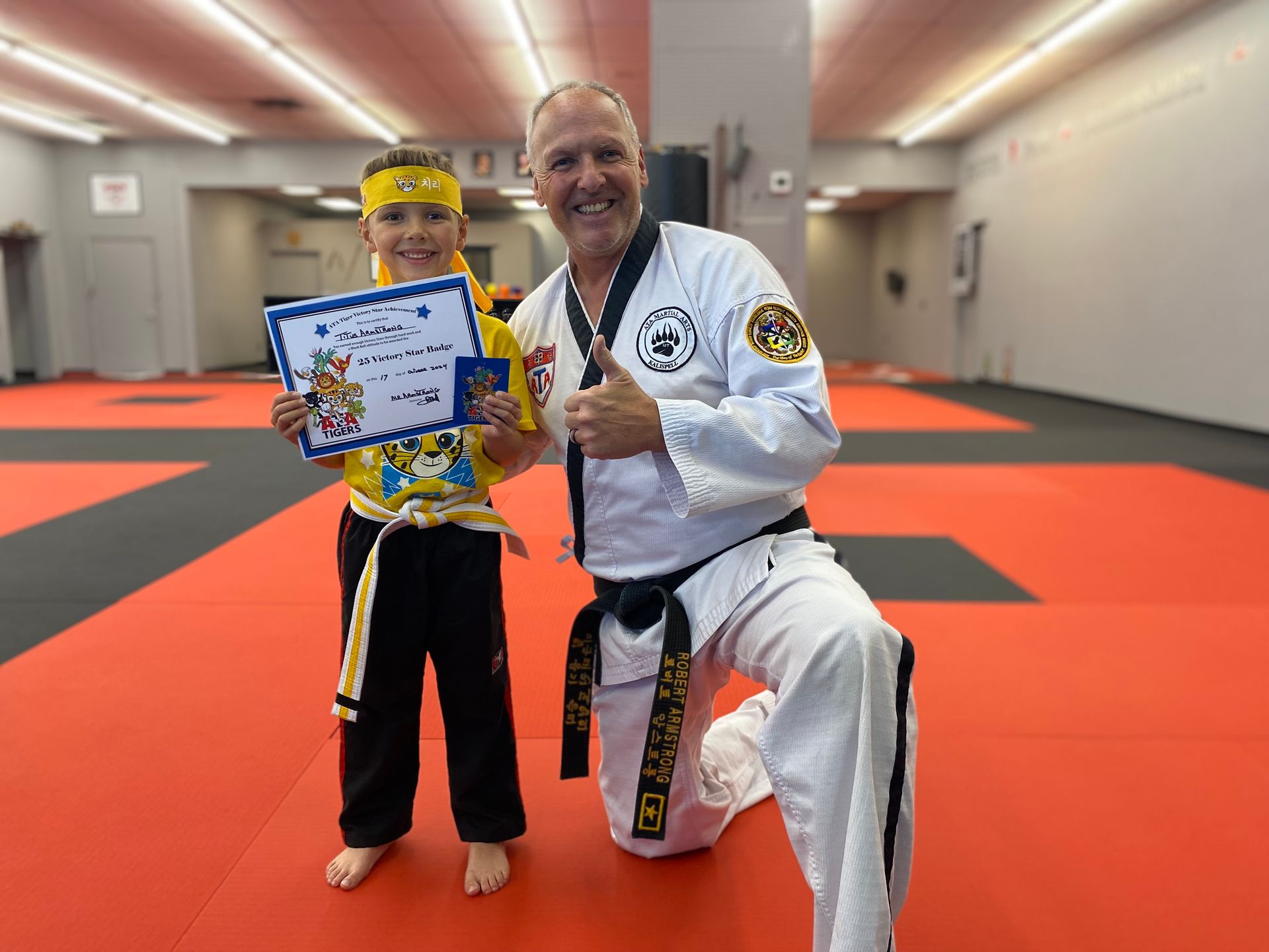 Boy in yellow belt and martial arts instructor kneel on a red mat, smiling and holding a certificate.