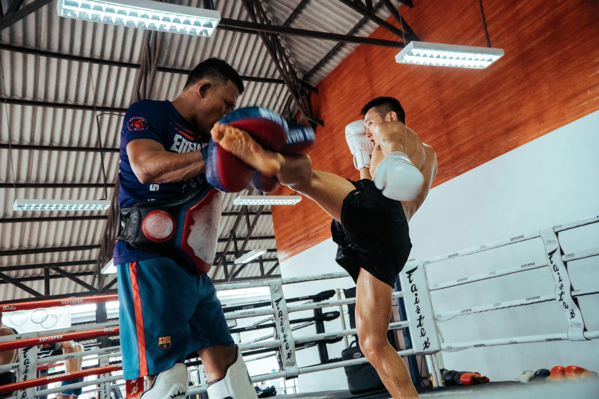 Man kicking pads held by another man in a boxing ring, practicing martial arts.